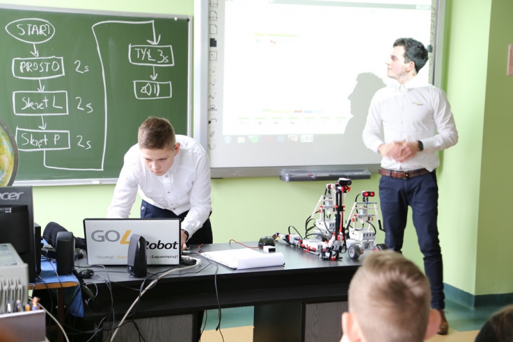 Prezentacja w sali lekcyjnej. Mężczyzna stoi przy tablicy multimedialnej, a jeden z uczniów pracuje przy komputerze.